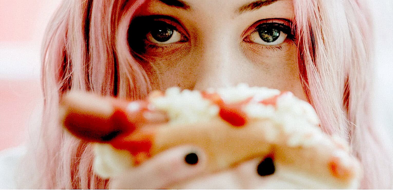 Delicious slice of pizza held close to a woman's face with pink hair, showcasing a craving for comfort food and vibrant personality, ideal for food and lifestyle content.
