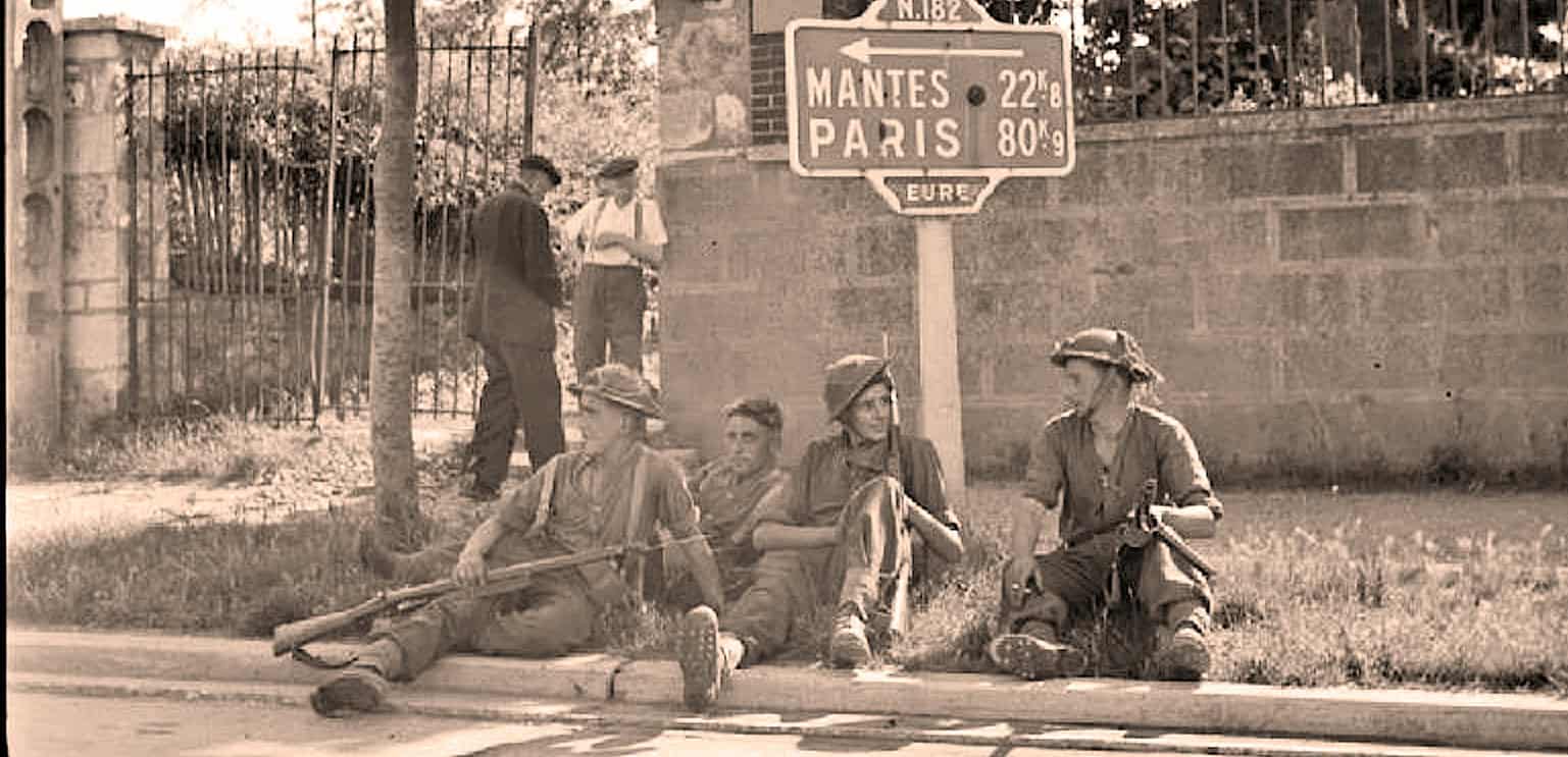 Soldiers resting at a European street corner during wartime, with a sign pointing to Mantes and Paris, capturing a historical moment from the past.