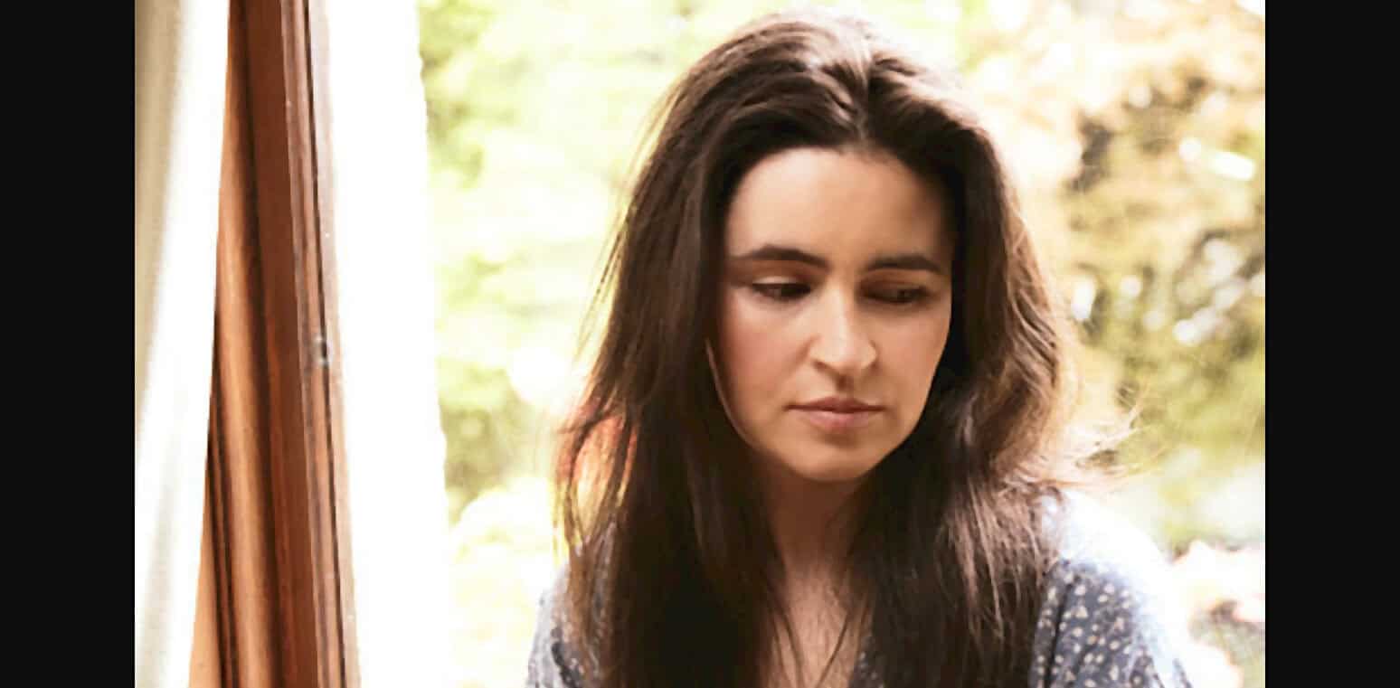 Young woman with long brown hair looking down by an open window on a sunny day, portrait of a girl lost in thought, nostalgic mood, natural light, soft focus.