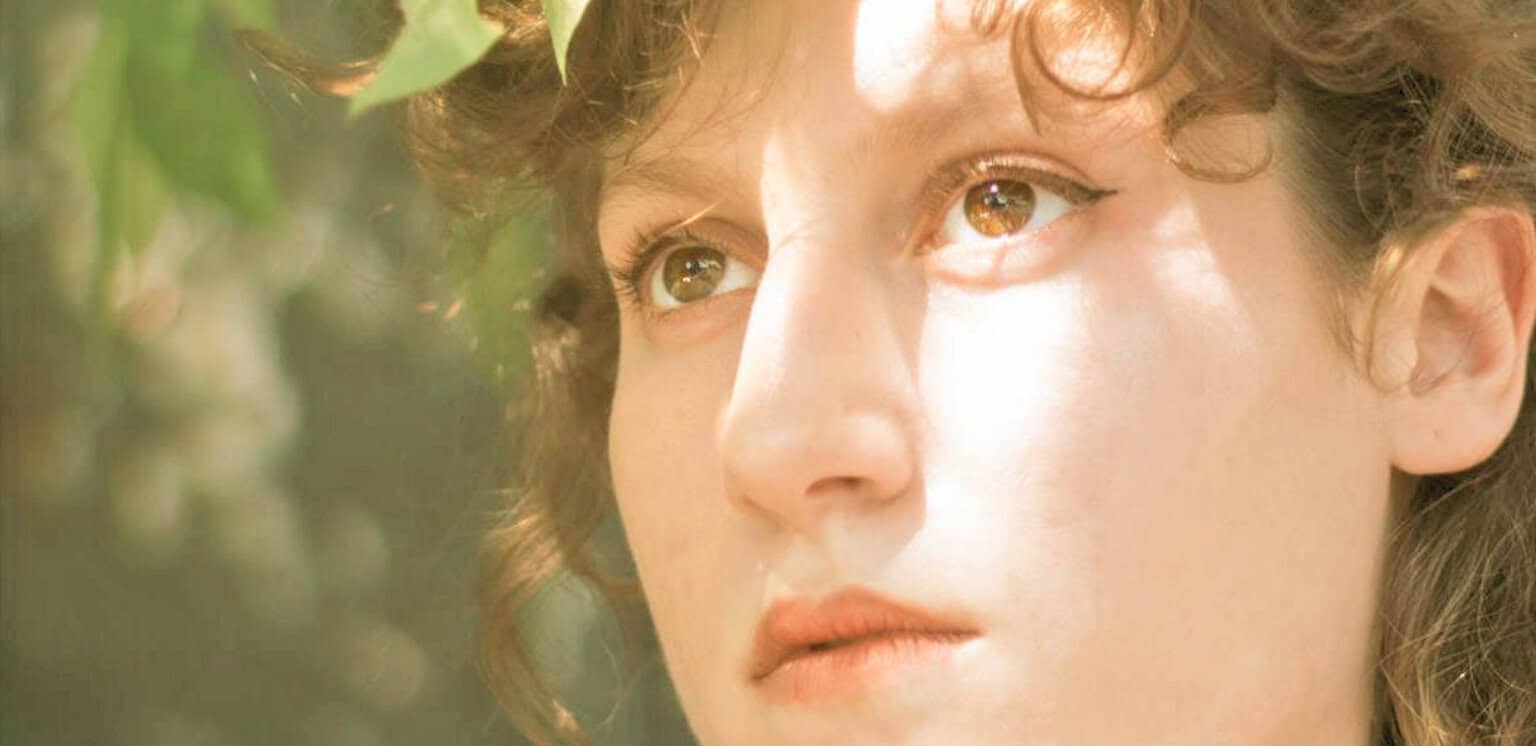 Close-up of a young woman's face with curly hair, soft natural sunlight, outdoor setting, capturing a thoughtful expression, emphasizing her brown eyes and delicate features, related to news, history, and music archives.