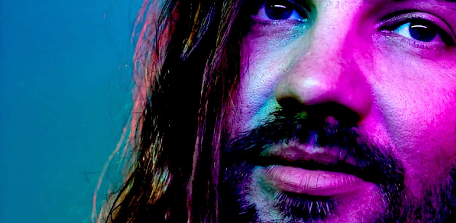 Close-up of a man's face with long hair and a beard, illuminated in vibrant neon pink, purple, and blue, emphasizing expressive eyes and deep facial features, representing music, news, and history archive themes.