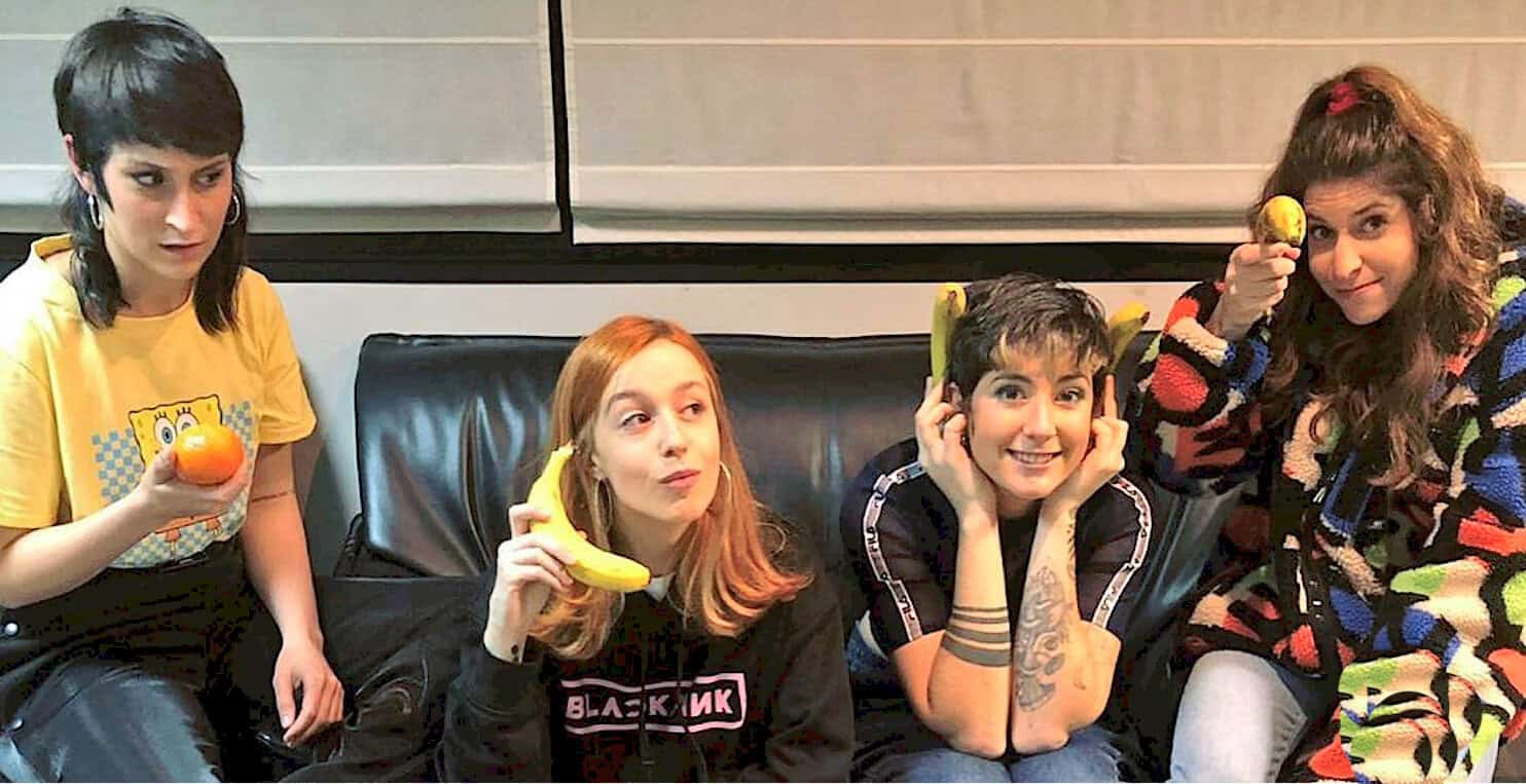 Fruit-themed group of young women with colorful accessories, holding fruit props, casual fashion, playful mood, trendy hairstyles, in a modern indoor setting, emphasizing fun and friendship.
