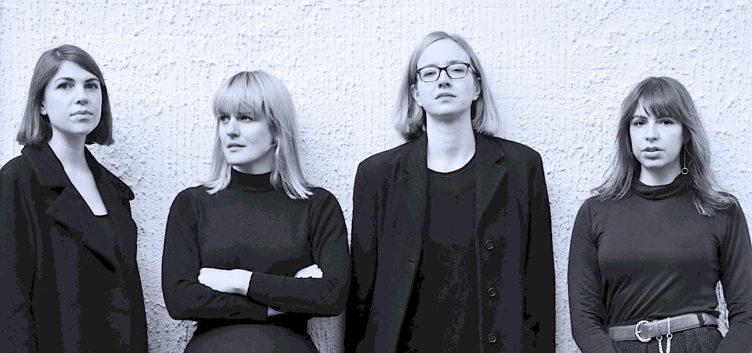 Black and white photo of four women band members standing against textured wall, showcasing music history, vintage news archive, and cultural sound recordings on Past Daily site.