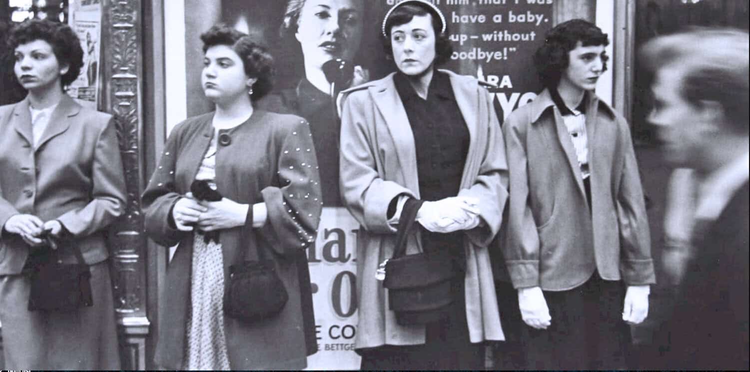 Vintage women waiting outside a newsstand, black and white photo, early 20th-century street scene, historical news, women's fashion, urban life, archival photograph for historical news and music archives.