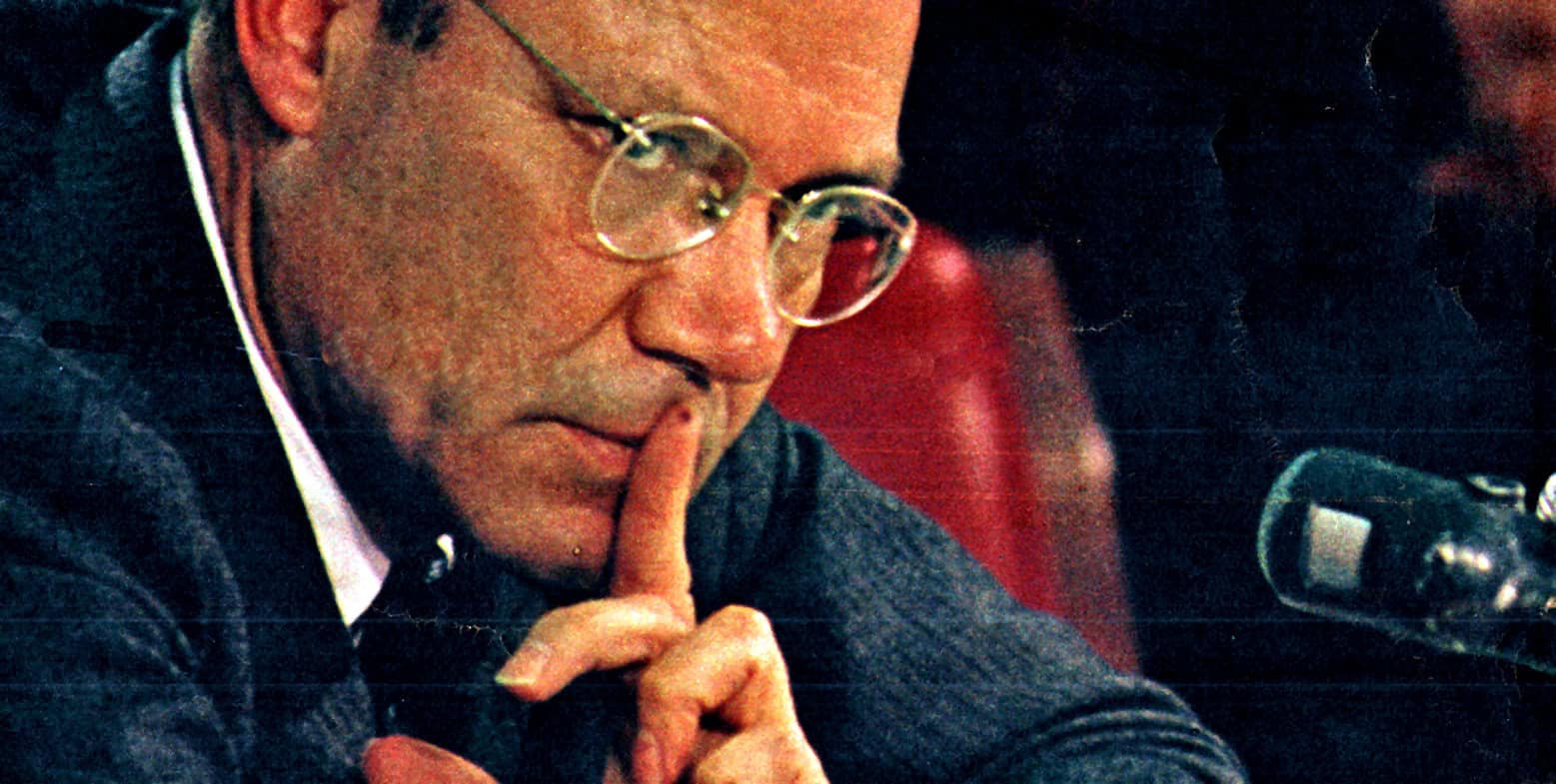 Thoughtful man in glasses with finger to lips, symbolizing silence or reflection, speaking at a news or history event, captured for a sound archive of news, history, and music.
