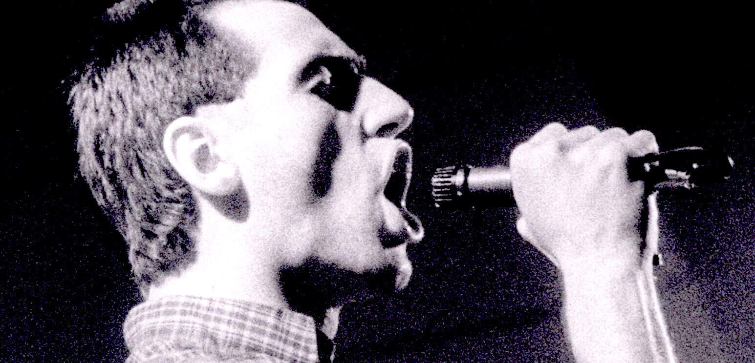 Black and white photo of a singer with short hair and sunglasses passionately singing into a microphone, highlighting vintage music and radio broadcast history.
