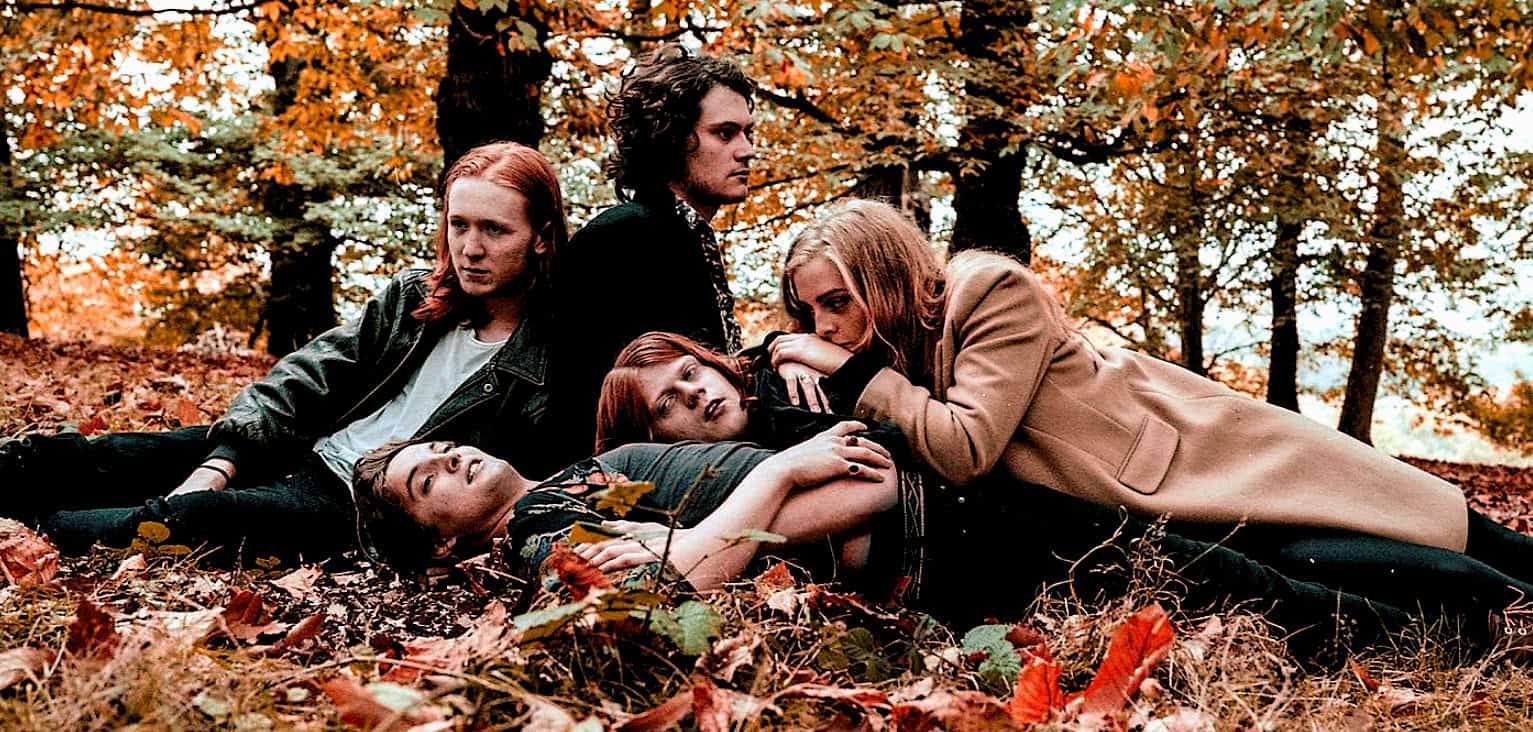 Vampire Academy cast lying on autumn leaves in a wooded setting during a fall photoshoot, showcasing gothic fashion and mysterious expressions.