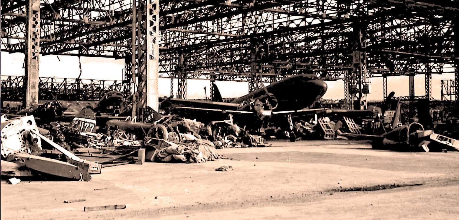 Destroyed aircraft and wreckage inside an aircraft boneyard, showcasing abandoned military planes with a metal framework overhead; historical aviation site.