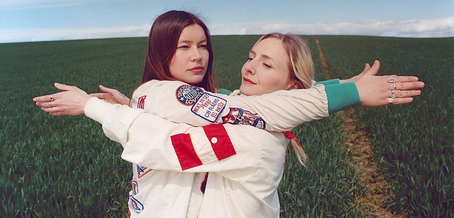 Vintage women in astronaut jackets embracing outdoors, representing music and history, with green field background.