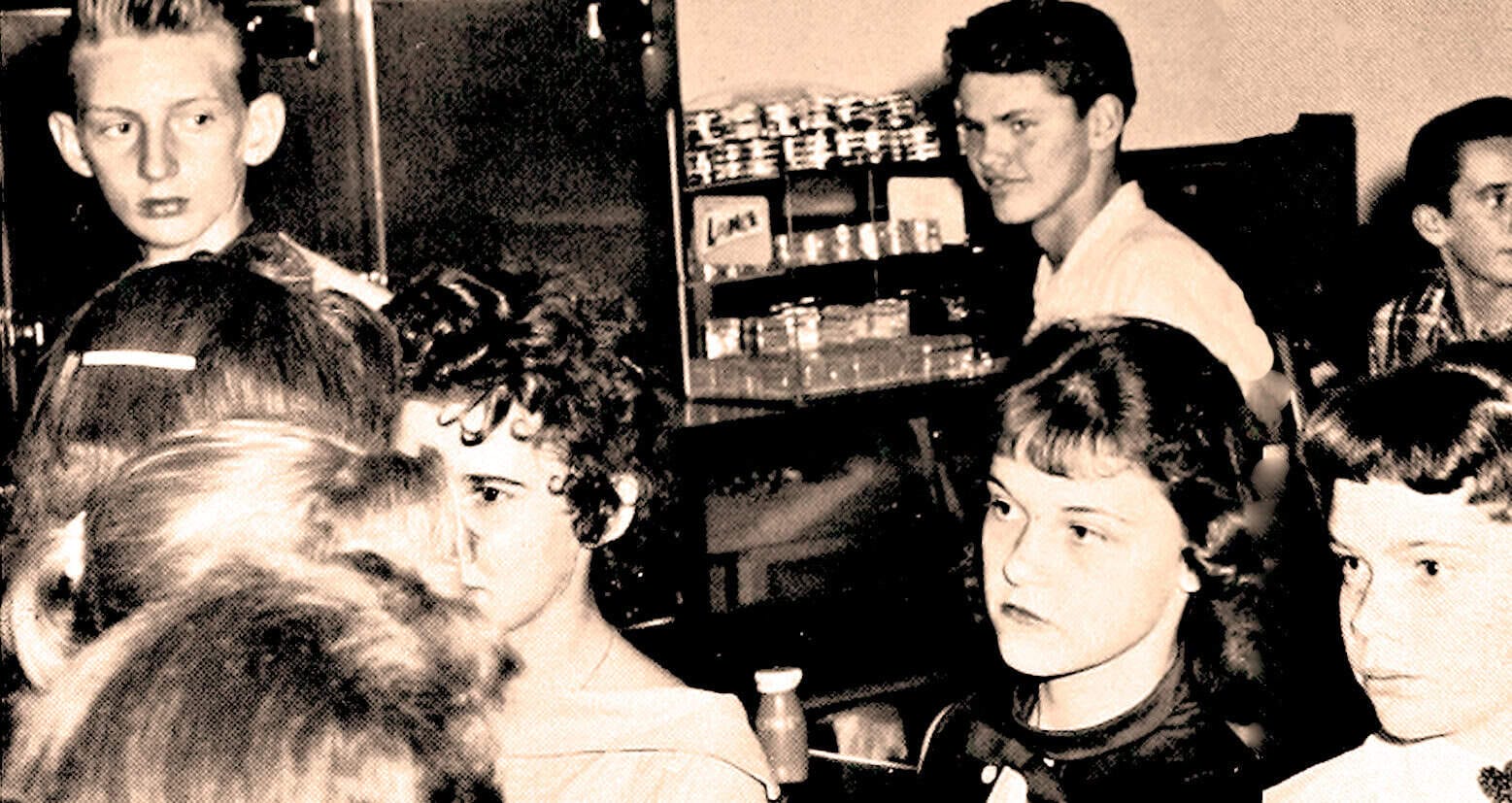 Vintage black-and-white photo of teenagers socializing in a 1950s or 1960s setting, capturing youth culture, fashion, and music influence from that era.