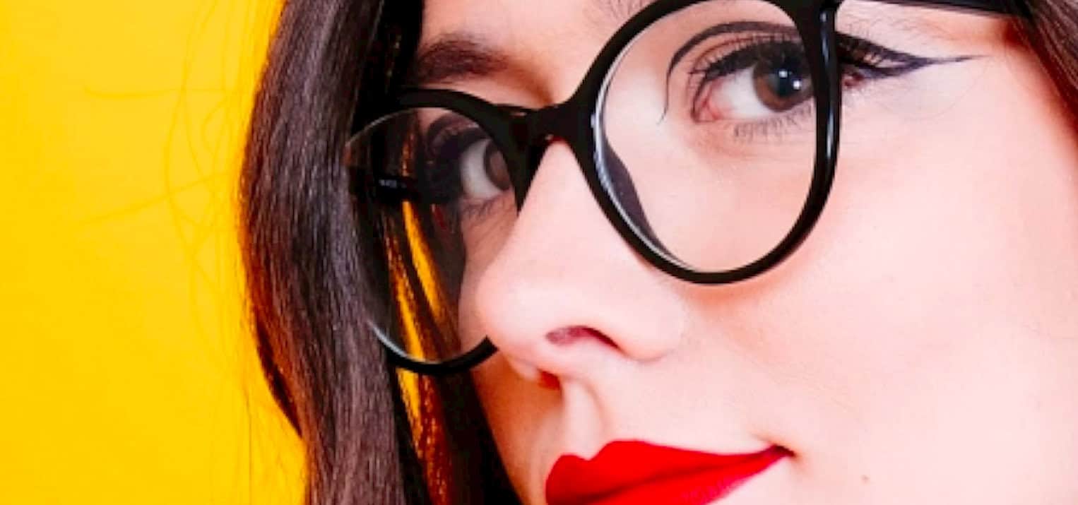 Close-up of a woman with dark hair, wearing round black glasses and red lipstick, representing a vintage or retro style inspired by historical news and musical archives.