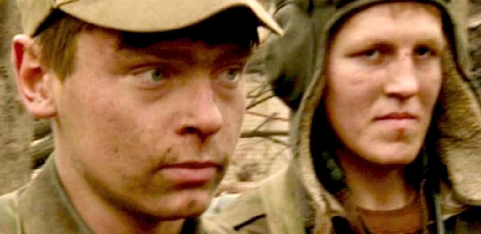 A vintage scene from the classic movie "The Battle of Algiers," showing two young resistance fighters with intense expressions, wearing military attire and hoods in a historical urban setting.