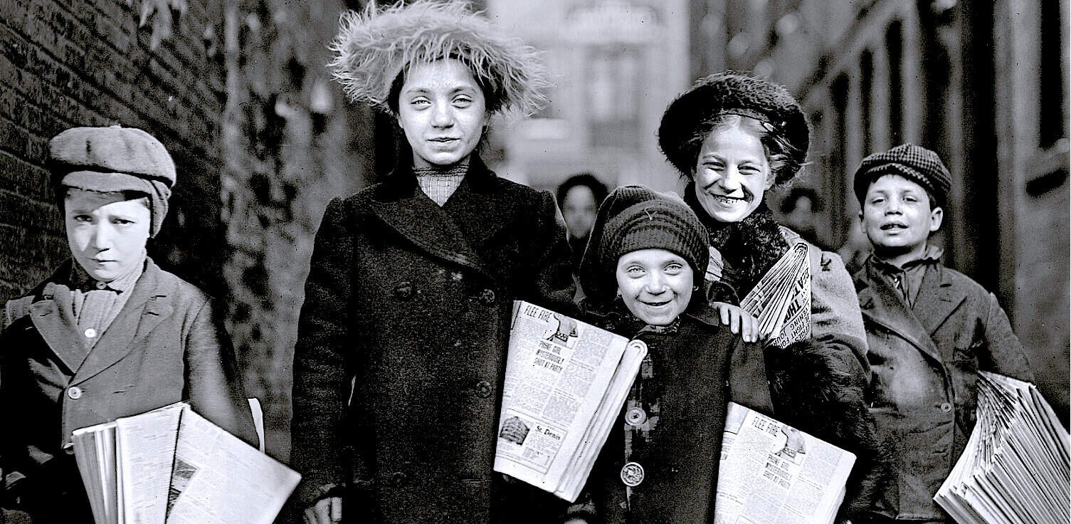 Child street vendors holding newspapers in a historical urban setting, representing news distribution and street commerce. Vintage black-and-white photo showcasing early 20th-century street life.