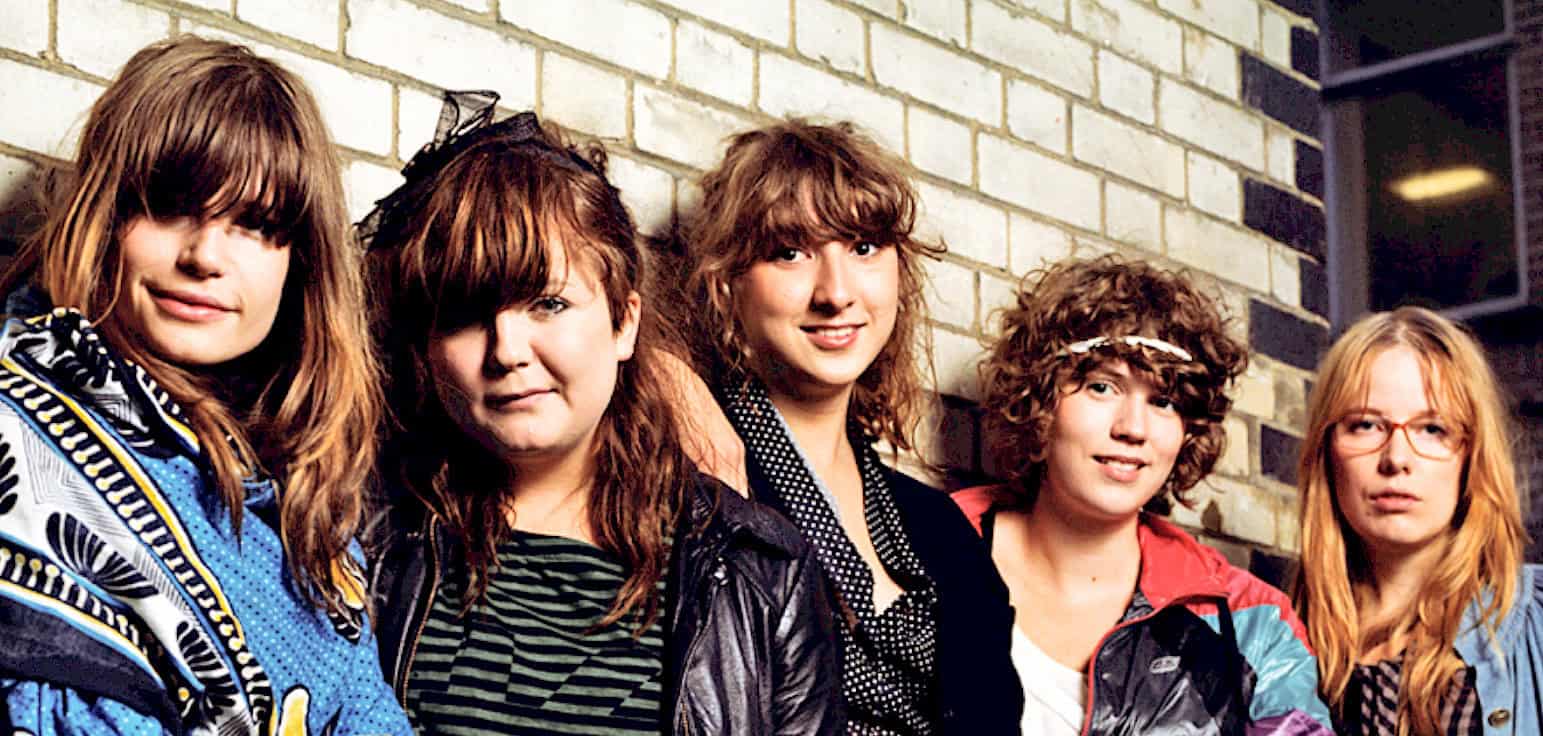 Teen girl band, indie rock girls, trendy women with curly and straight hair, casual fashion, urban brick wall backdrop for music or youth culture photography, Past Daily news archive, history, music, news recordings.