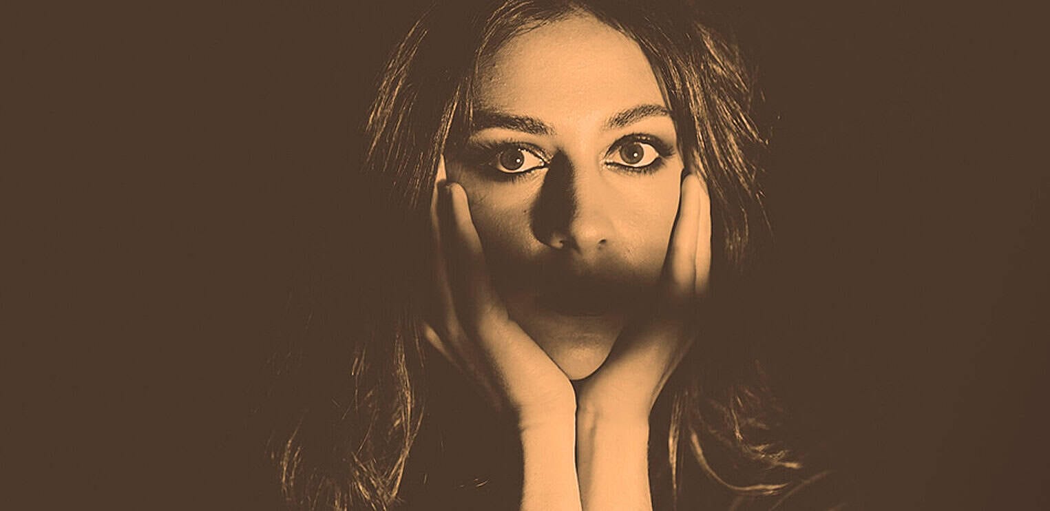 Close-up portrait of a woman with red hair and intense eyes, holding her face with both hands, representing history, music, and news archives at Past Daily.