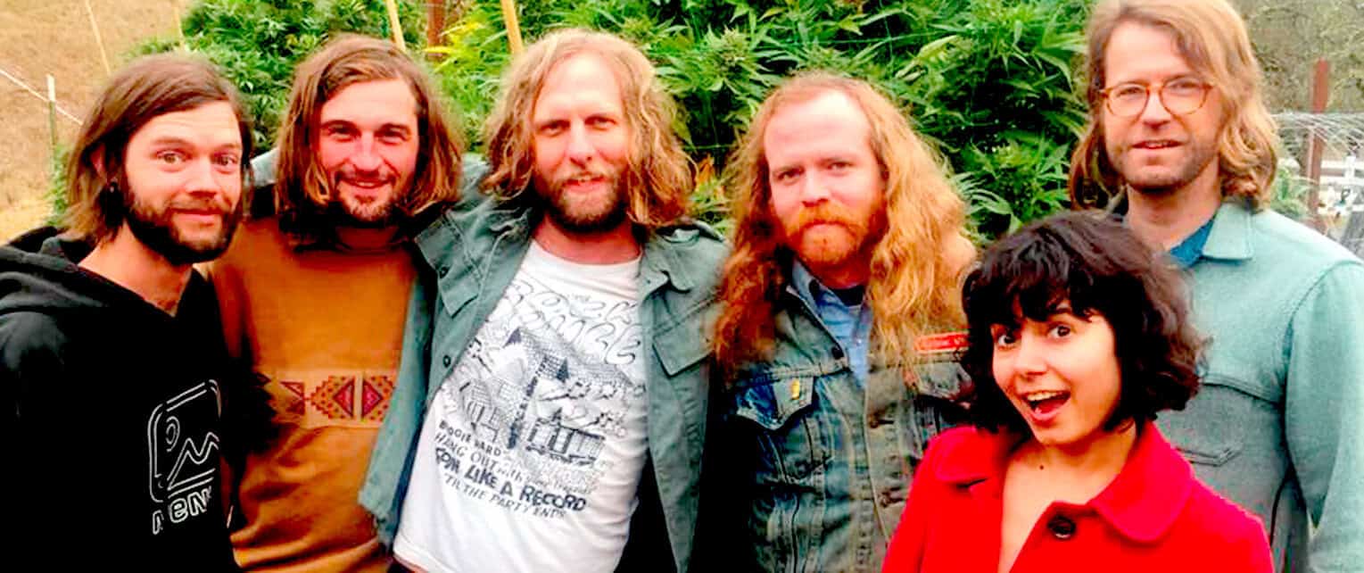 Group of five diverse young adults outdoors, enjoying a casual moment, surrounded by lush green plants, representing community, friendship, and upbeat lifestyle themes for news, music, and history archives.