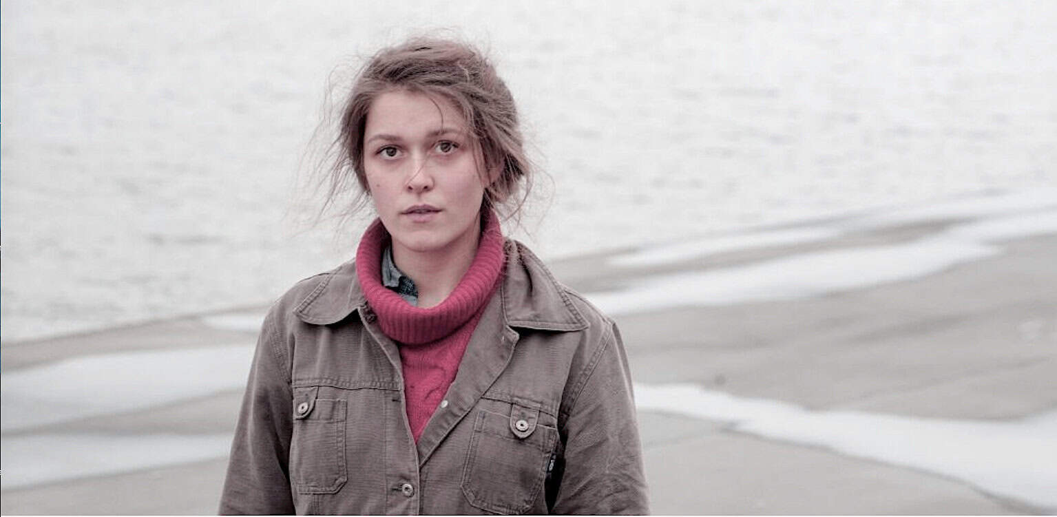 Naked woman by a body of water with a pink turtleneck and gray jacket standing on a beach, sound archive of news, history, and music, emotional portrait from Past Daily: A Sound Archive of News, History And Music.