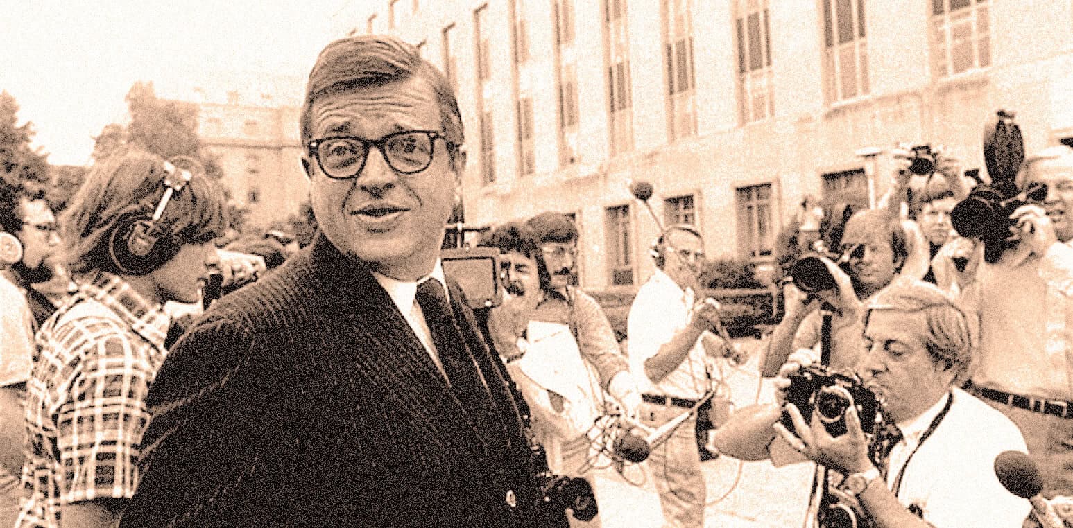 Photograph of a vintage news reporter surrounded by journalists and cameramen outside a historic building, capturing a moment of broadcast journalism from the mid-20th century.