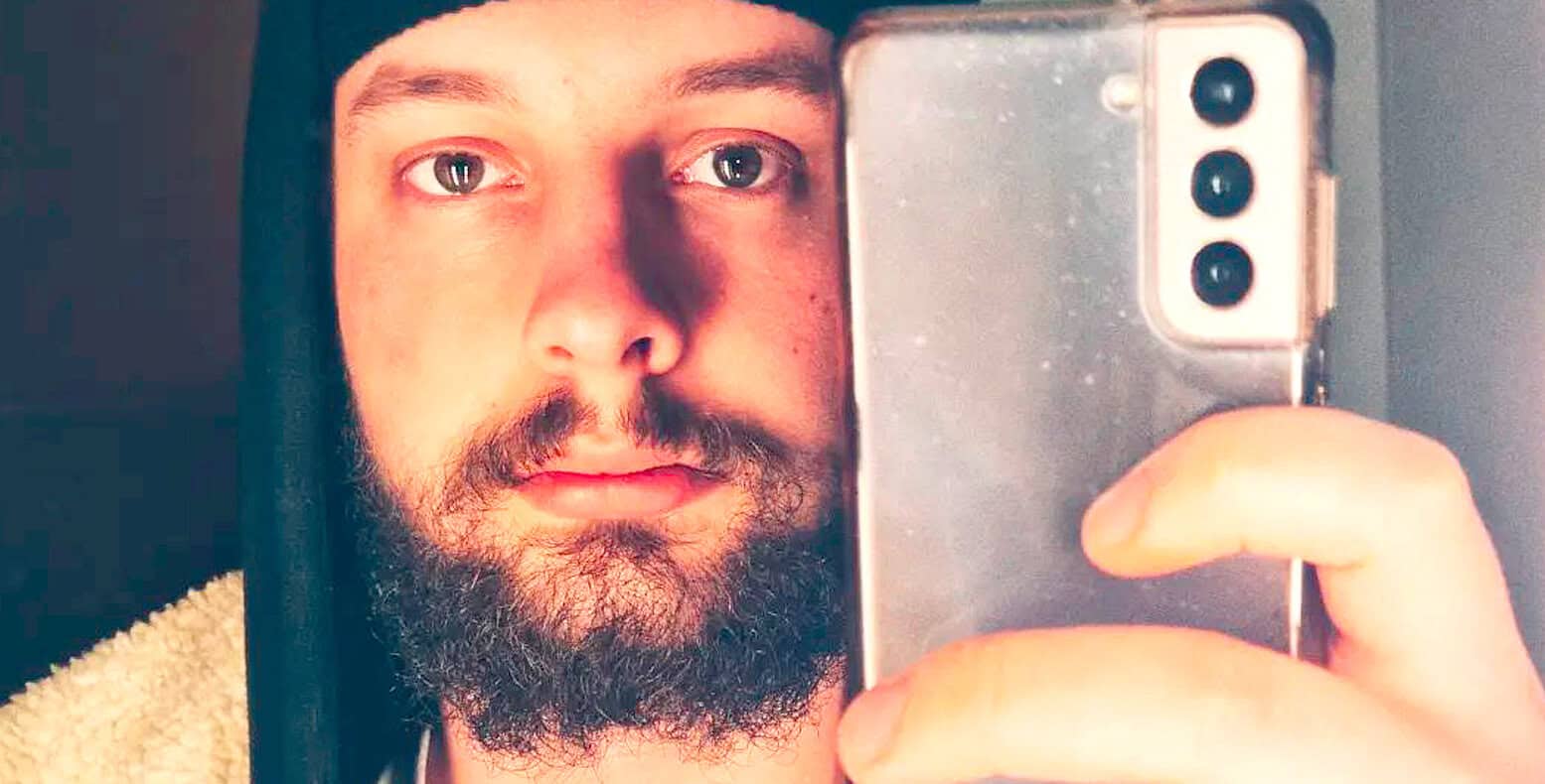 Close-up of a man taking a selfie with a smartphone, featuring a beard, blue eyes, and casual attire, highlighting modern technology and selfie culture.