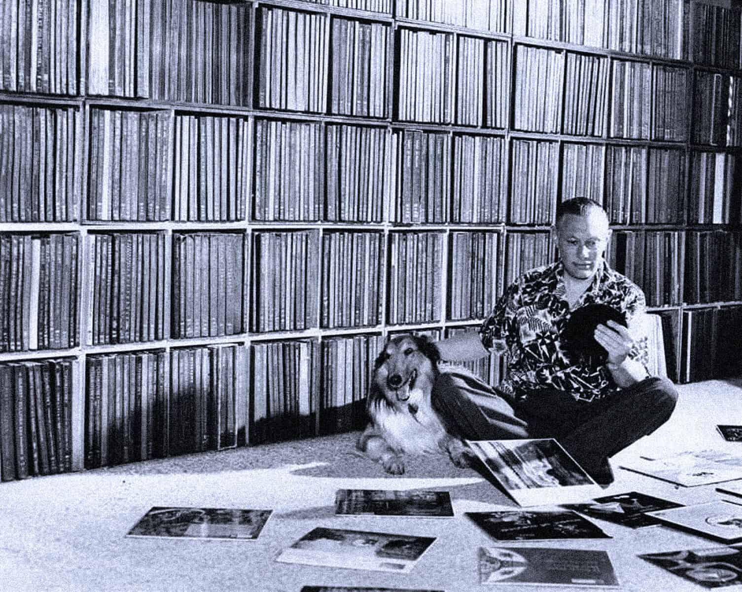 Vintage photograph of a person with a dog sitting on the floor amidst vinyl records, with a large wall of categorized vinyl records behind them, illustrating a sound archive, music collection, and historical record storage.