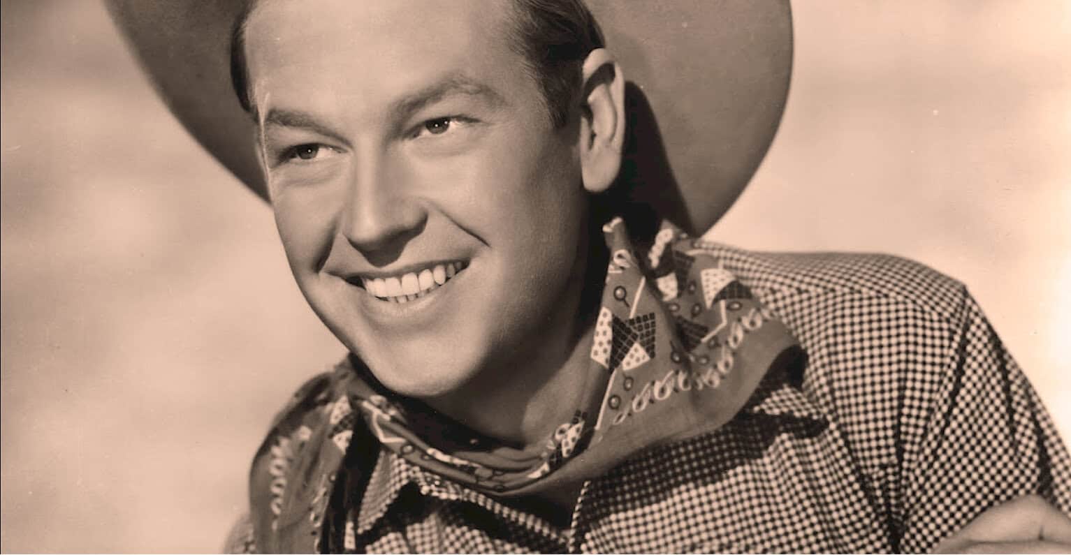 Vintage black and white portrait of a smiling man in cowboy attire, featuring a wide-brimmed hat and patterned neckerchief, perfect for history and music archive images.