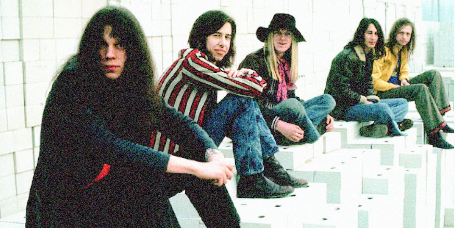 Colorful 1990s grunge band members sitting on white stairs, casual style, music scene, alternative rock, youth culture, vintage concert vibe, street style, band photograph, edgy fashion, music history.