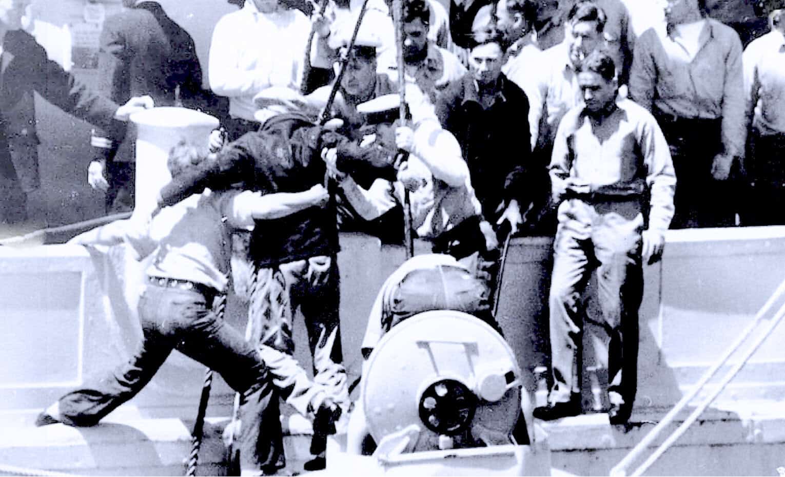A historical black and white photo depicting a police officer using a water cannon during a protest, with crowds of onlookers and protesters in the background, capturing moments of civil unrest and public demonstrations.