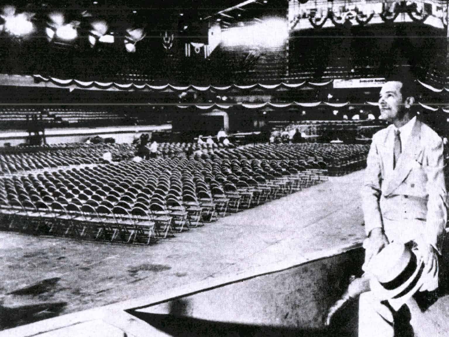 Vintage photo of a man in a suit holding a hat, standing in a large hall filled with empty chairs, stage curtains, and theatrical lighting, representing historical news, music, and event archives.