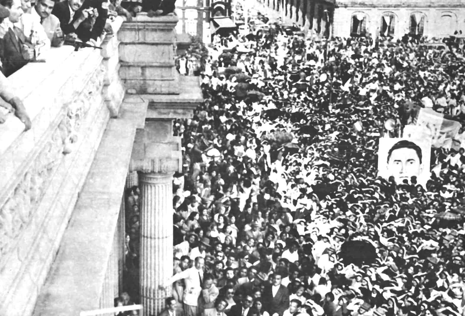 Crowd gathered at a political rally or protest, with a large portrait of a man holding a microphone displayed prominently in the background, symbolizing social or political activism.