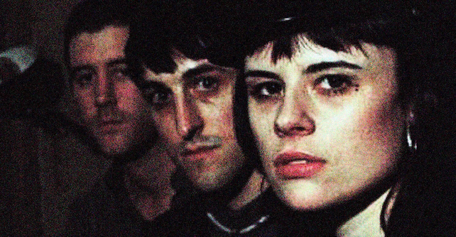Woman with short dark hair and piercing eyes, behind a group of people appear to be in a cinematic scene, dark background, intense expression, strong visual storytelling, related to news, history, or music archives.