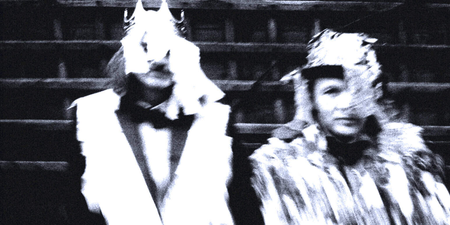 Black and white photo of two women in vintage clothing, one with a bow tie, sitting on theater stairs, representing historical news, music, and cultural archives from Past Daily.