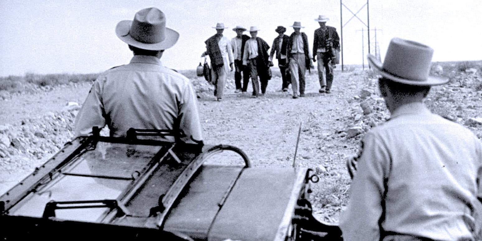 Vintage black and white photo of men walking down a dusty road with law enforcement officers in the foreground. Historical music, news archive, and history audio recordings available at Past Daily.