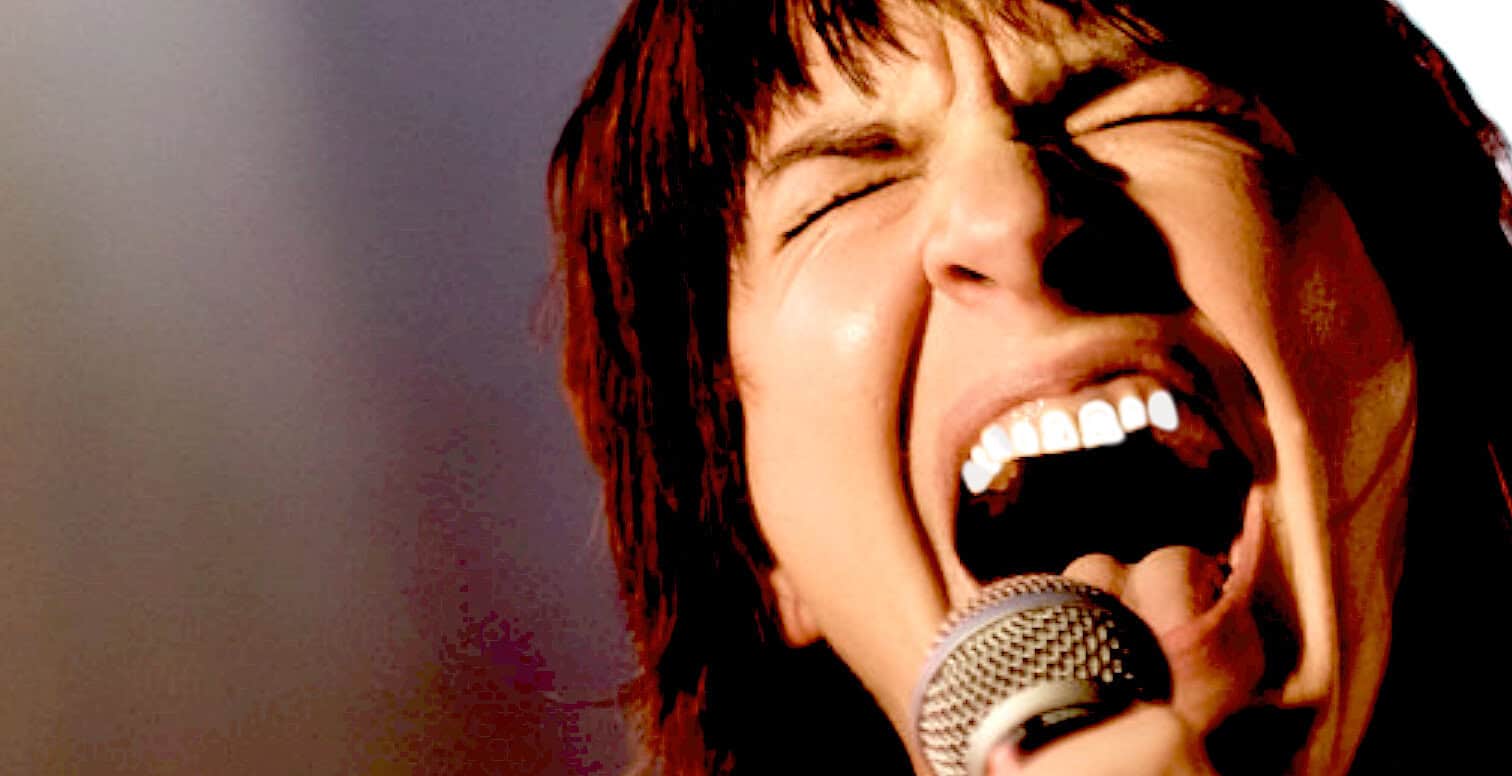 Passionate female singer singing into microphone, expressing emotion with open mouth, energetic music performance, rock and pop genre, vibrant stage presence, music archive, news, history, and music history imagery.