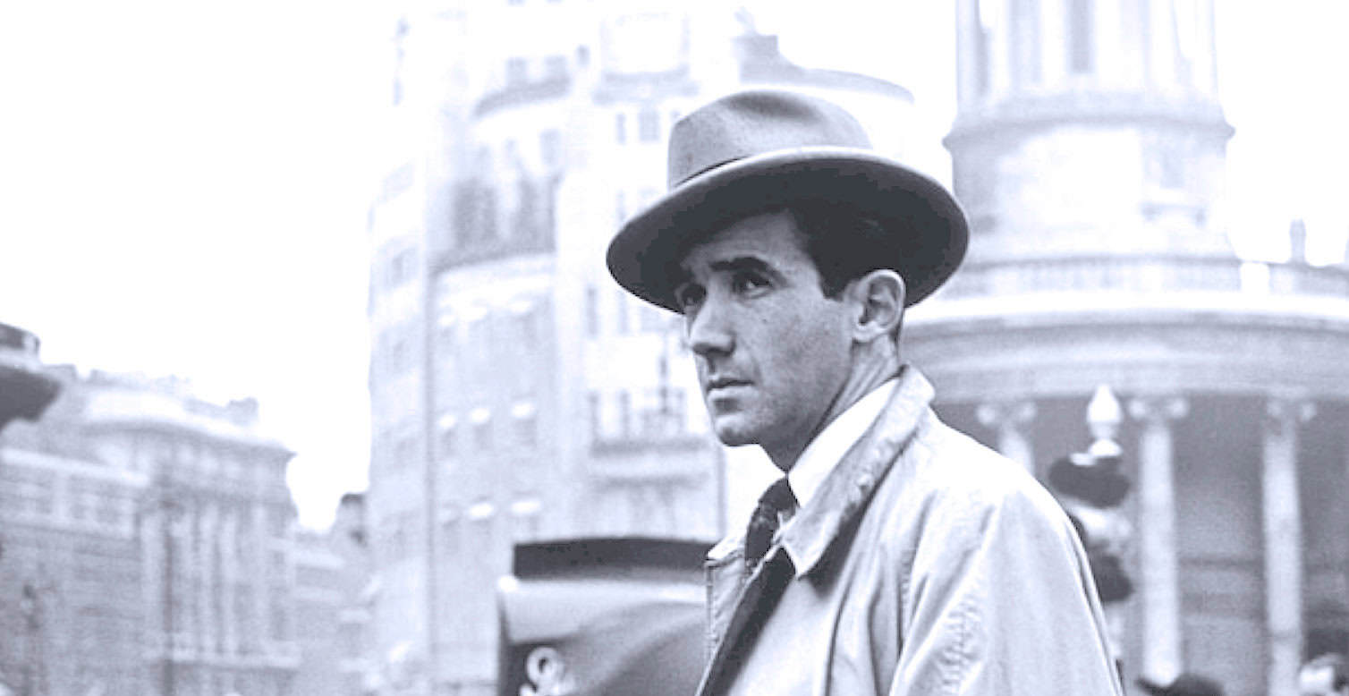 Man in trench coat and fedora hat looking sideways on busy city street, black and white photo.
