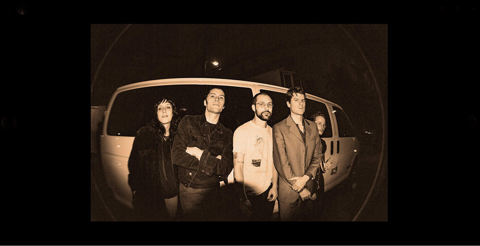 Mellow yellow band standing in front of a van at night, sepia toned image, indie rock music, band photo, retro style, music archive, historical music scene, artist promotion, past daily sound archive, music history exploration.