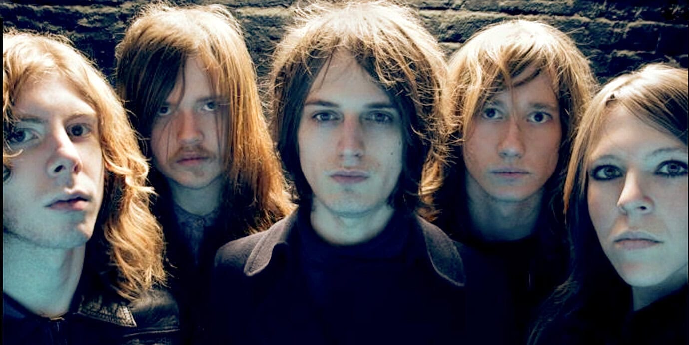 Vintage rock band with long hair and leather jackets, posing against a dark brick wall, representing music history and classic rock culture.