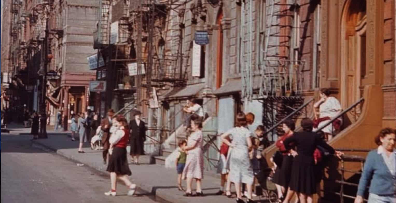 Historic city street scene with women and children waiting outside brownstone buildings during daytime, vintage urban atmosphere, mid-20th century, 1950s, city life, community, historical news footage, music archive focus.