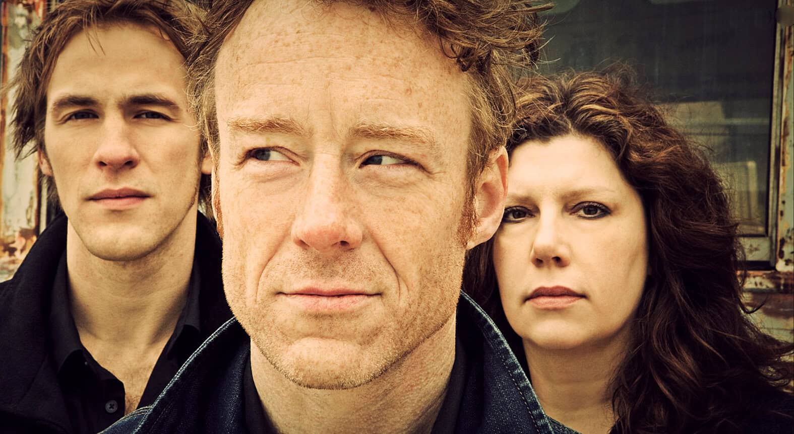 Close-up of a man with red hair and freckles, flanked by a young man and woman with intense expressions, set against an urban background; perfect for music, history, and news archive visuals.