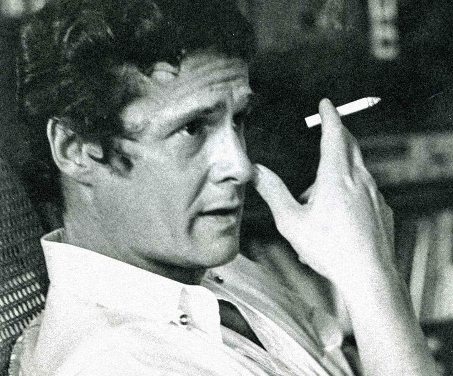 Vintage black-and-white photo of a man with dark, wavy hair, holding a cigarette near his face, reminiscing classic moments from music, news, and history archives.