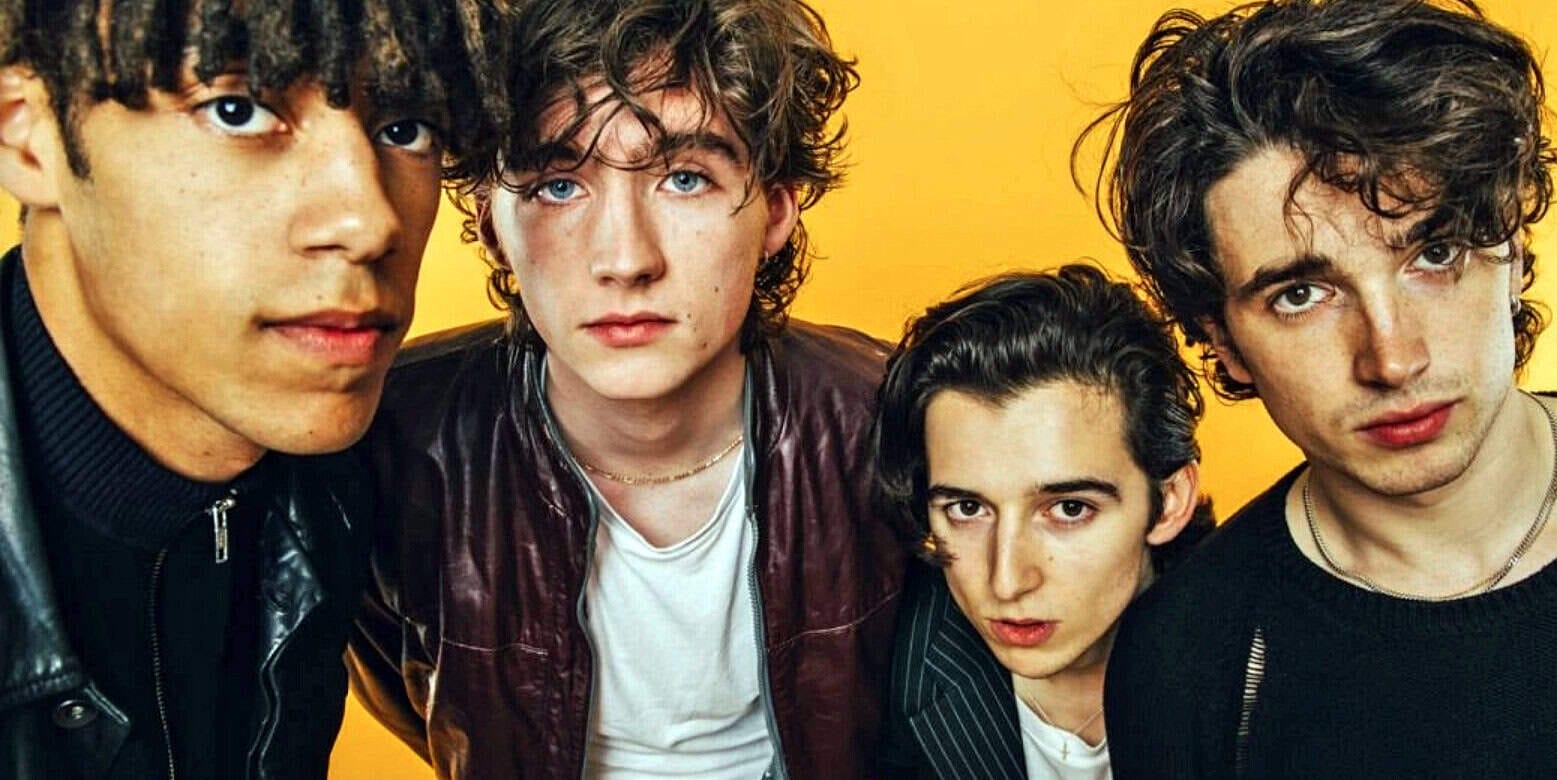 Youthful band members with curly and straight dark hair, wearing casual leather and striped clothing, posing against a vibrant yellow background.