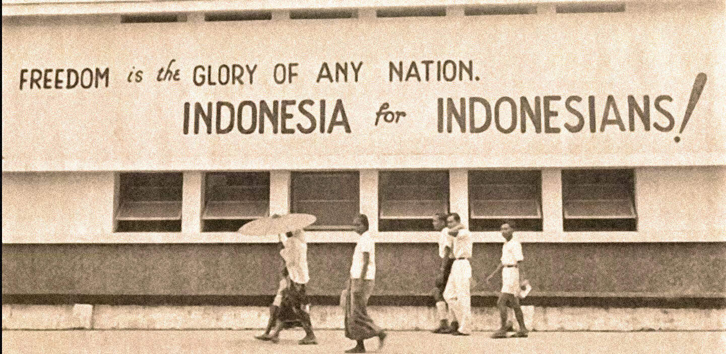Vintage black and white photo of people walking past a building with a large banner advocating Indonesian independence, capturing a historic moment in Indonesia's fight for freedom.