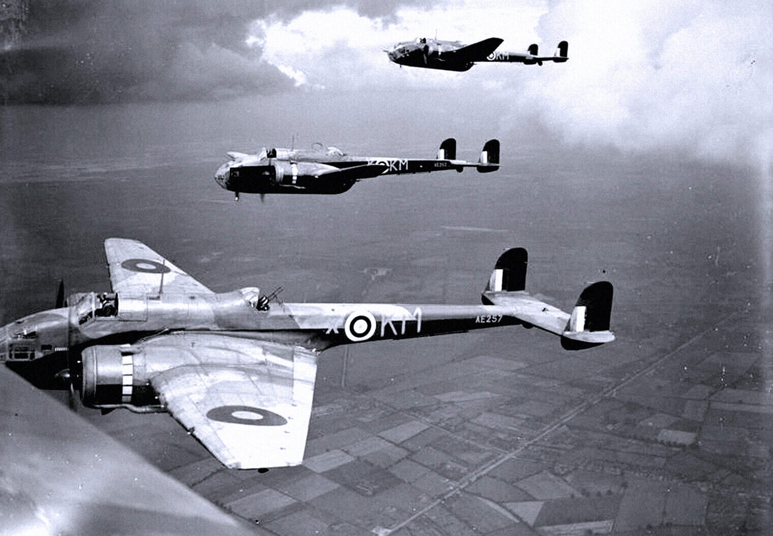 Vintage WWII-era British aircraft flying formation over farmland, black and white image, historic military aircraft, aviation history, iconic warplanes, air force heritage, past daily archive, military aviation.