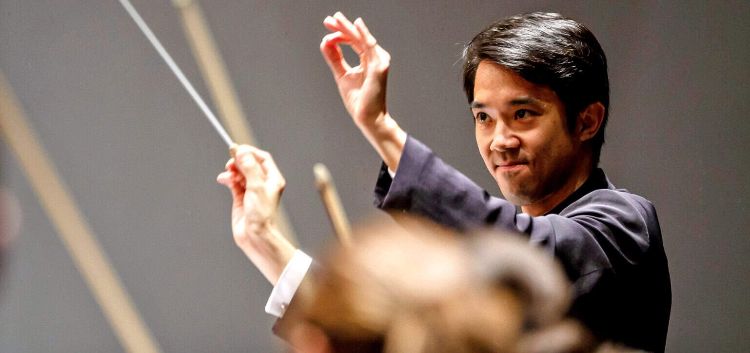 Orchestral conductor leading a symphony with baton, focused on music performance, in a concert hall.