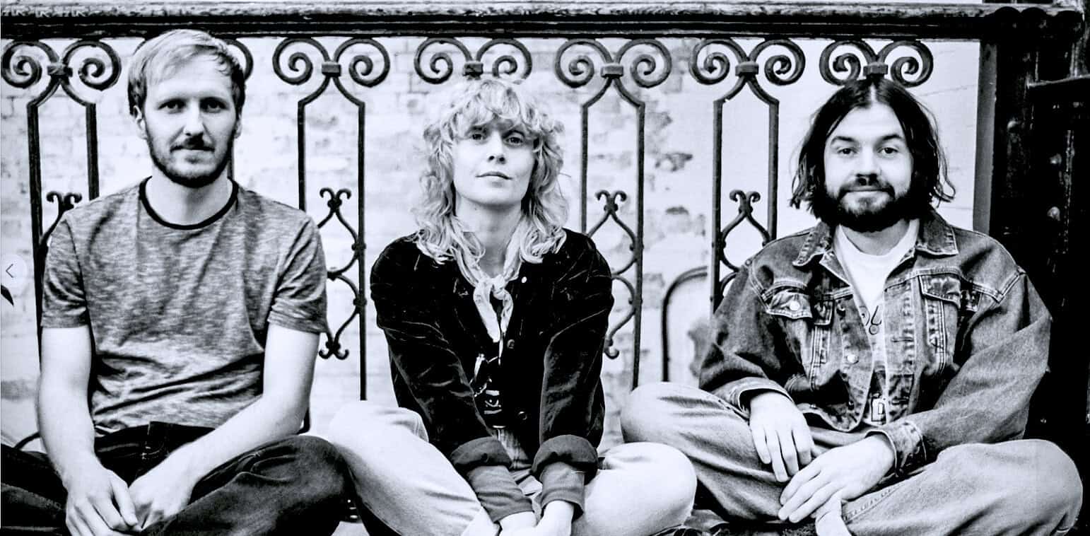 Bold black and white photo of three young musicians sitting against an ornate wrought iron fence, representing a classic image of music history, nostalgia, and artistic expression, from the Past Daily sound archive.