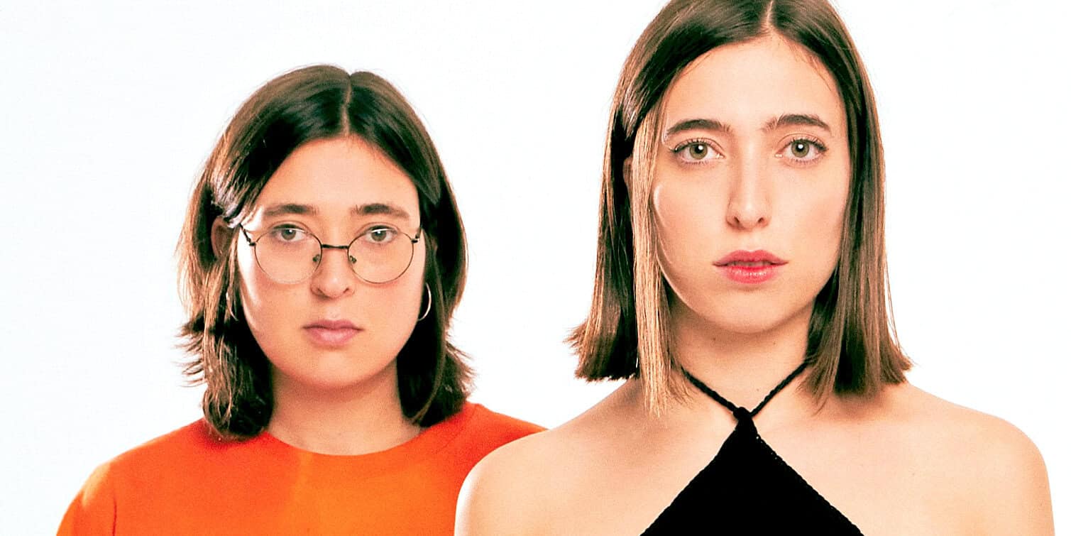 Close-up of two young women with shoulder-length hair, one wearing glasses and a red top, the other in a black top, representing modern youth culture, music, and fashion, with a white background.