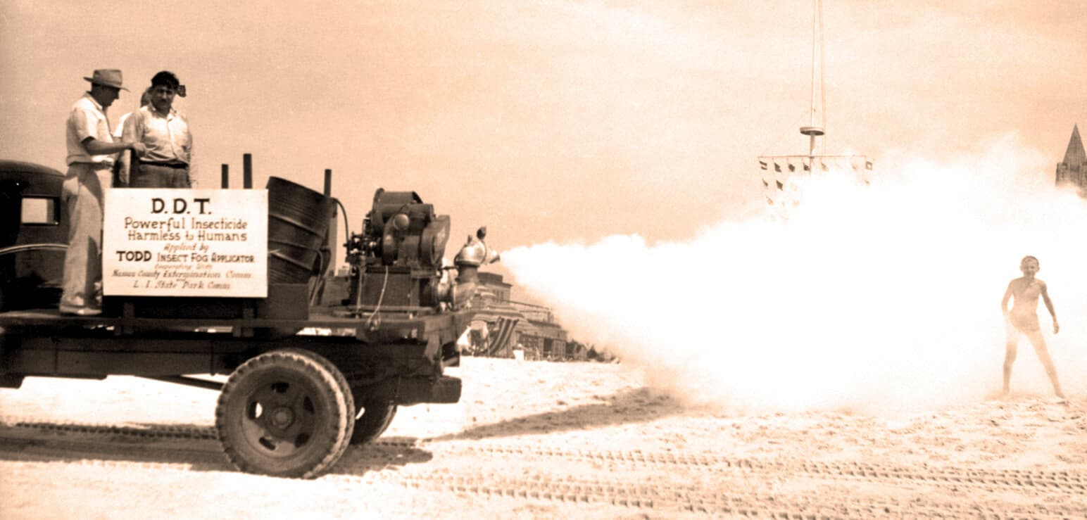 Insecticide spraying truck on sandy beach with a person walking, vintage black-and-white photo.