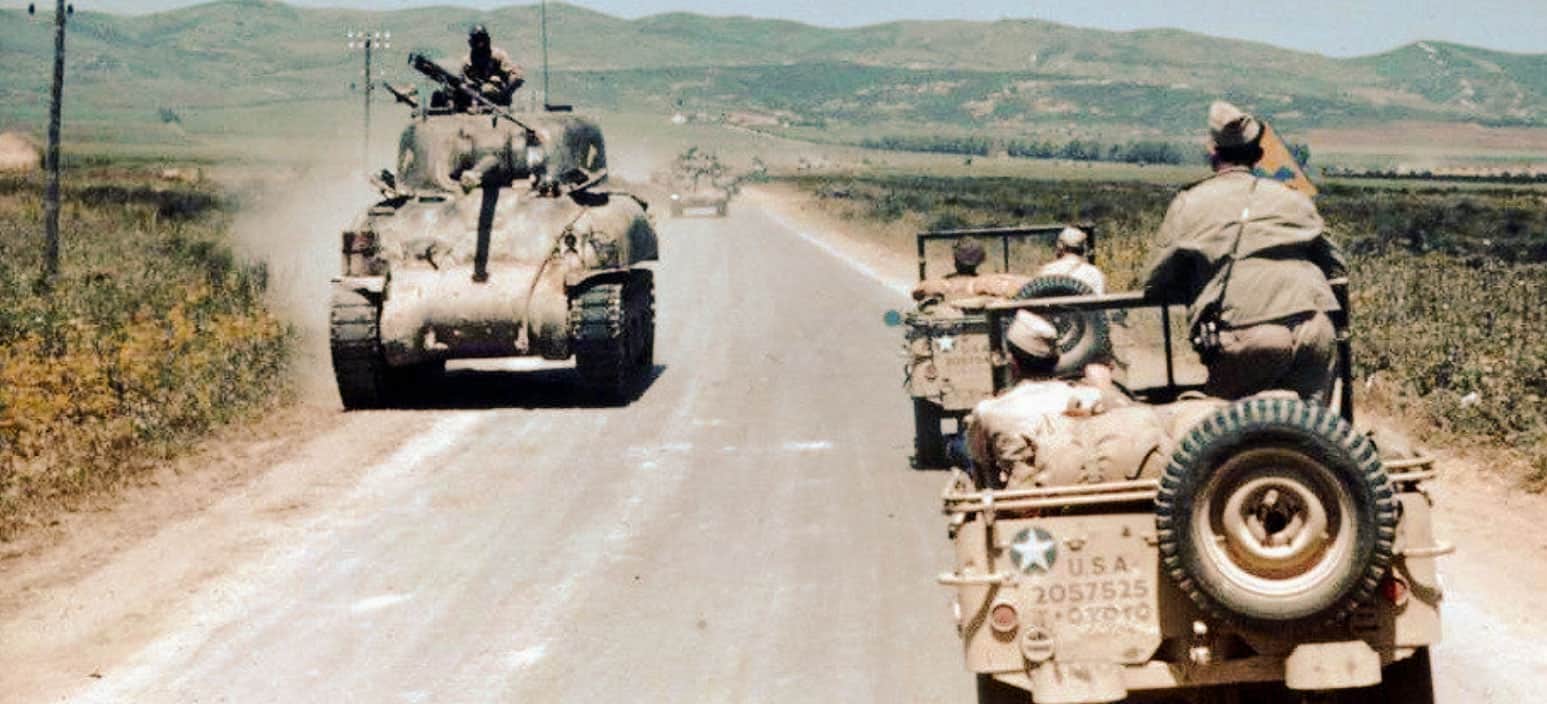 Military tanks and jeeps traveling on a rural road during a historical military convoy or operation, with mountains in the background and soldiers onboard, capturing a moment from wartime history, perfect for news and history archives.