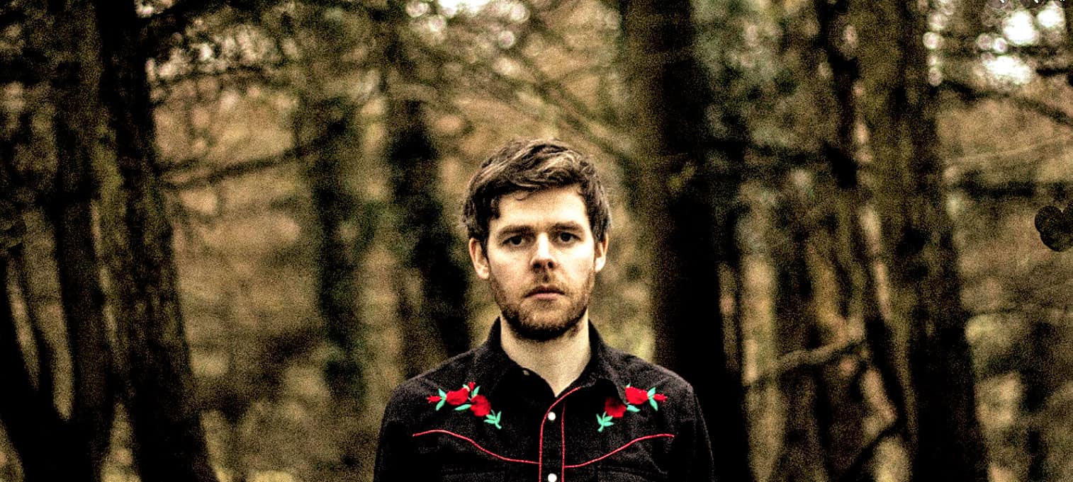 Solo male in black embroidered shirt standing in a forest with blurred trees in the background, capturing a moody and artistic image for music and history archives.