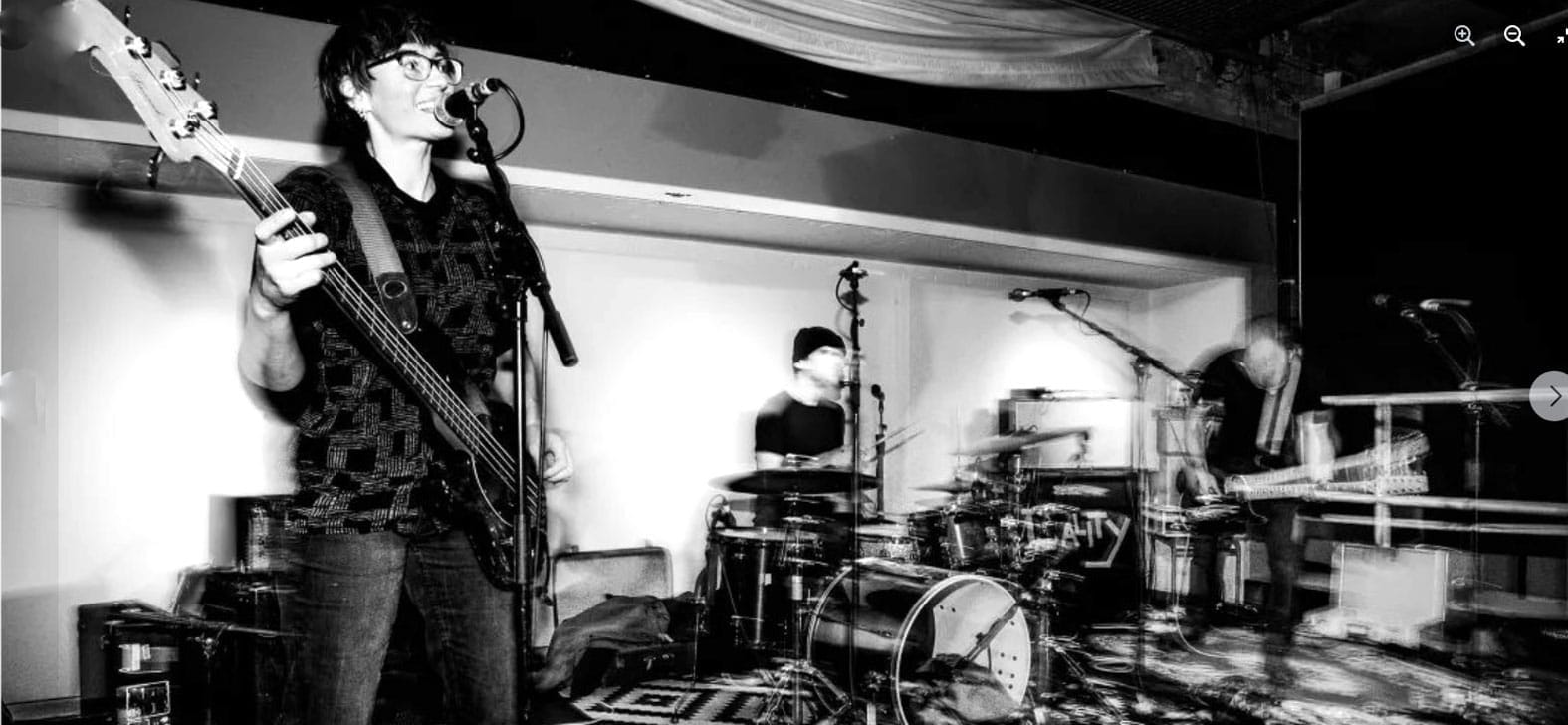 Woman playing bass guitar and singing at a music performance, live band on stage with drums and keyboard, black and white photo, music event, artist at Past Daily Sound Archive of news, history, and music.