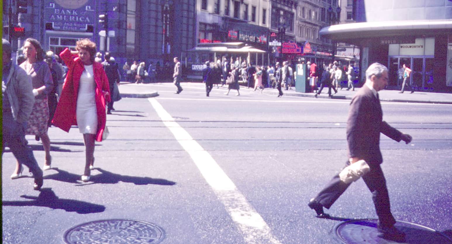 City street scene with pedestrians and shops, vintage city life photo, urban atmosphere, busy downtown, historic architecture, people crossing streets, 1960s fashion, commercial area, public transportation, bustling city environment, archive photo, old cityscape, nostalgic urban scene, news, history, and music documentation, Past Daily sound archive.